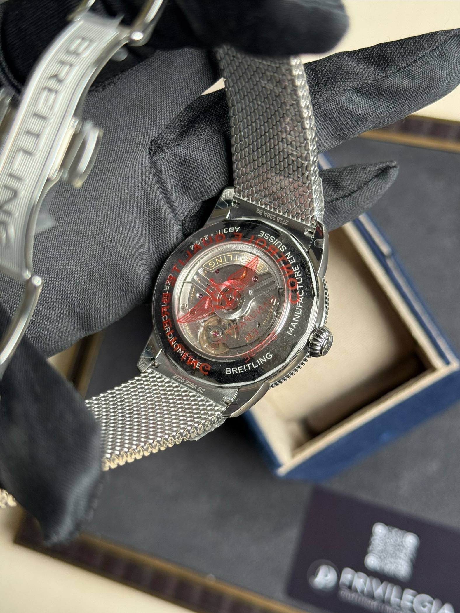 Back of a Breitling wristwatch with a metal mesh band resting on a gloved hand; you can see the red accent and engravings on the case back, including 'BREITLING' and 'MANUFACTURE'.