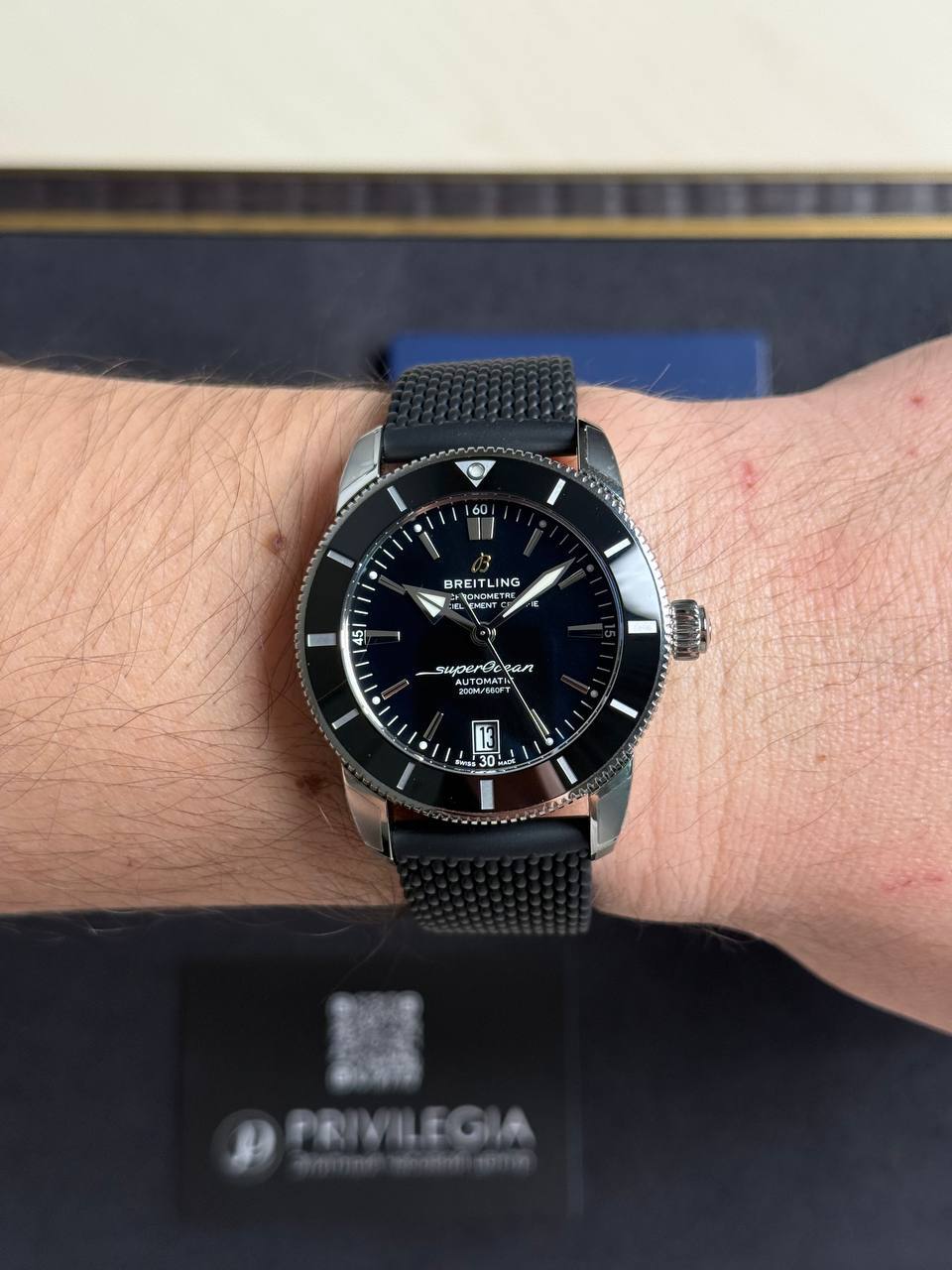 Close-up of a men's Breitling wristwatch with a black dial and metal mesh strap on a light-skinned wrist.