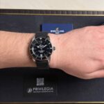 Breitling wristwatch with black dial and metal mesh band worn on the left wrist, blue Breitling card in the background.
