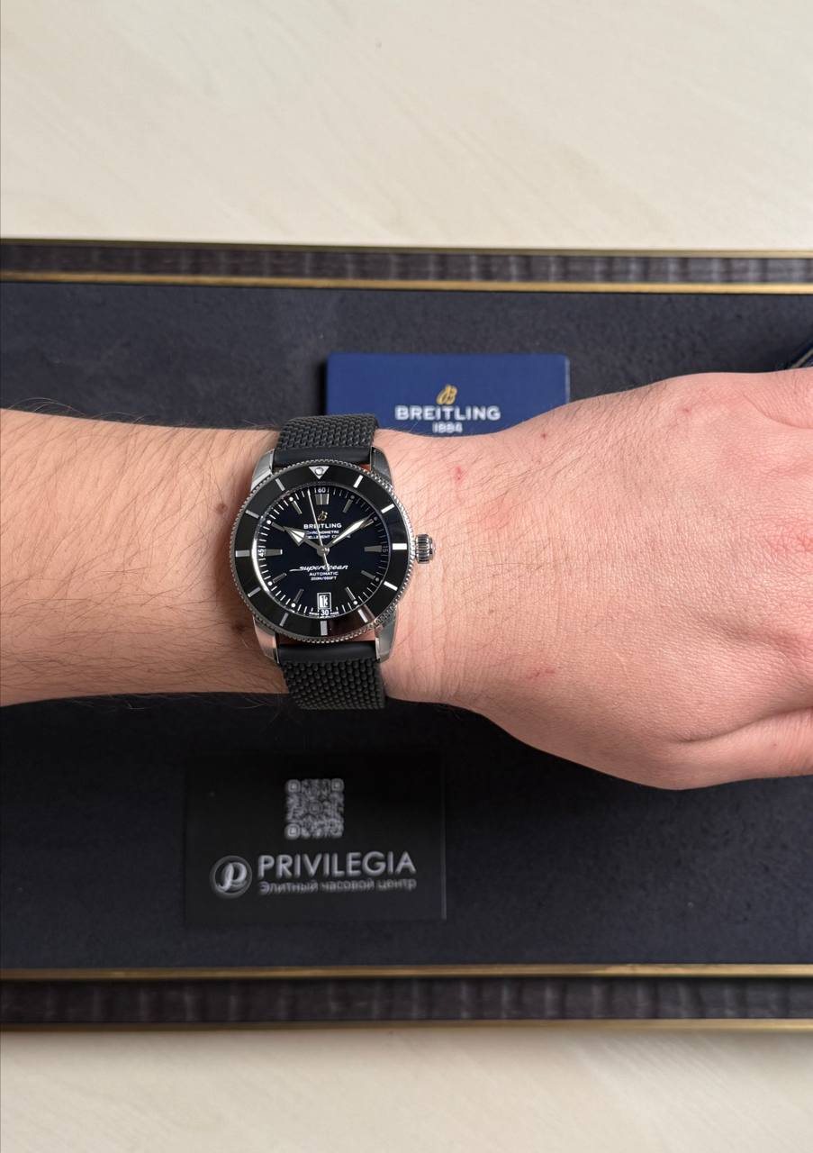 Breitling wristwatch with black dial and metal mesh band worn on the left wrist, blue Breitling card in the background.