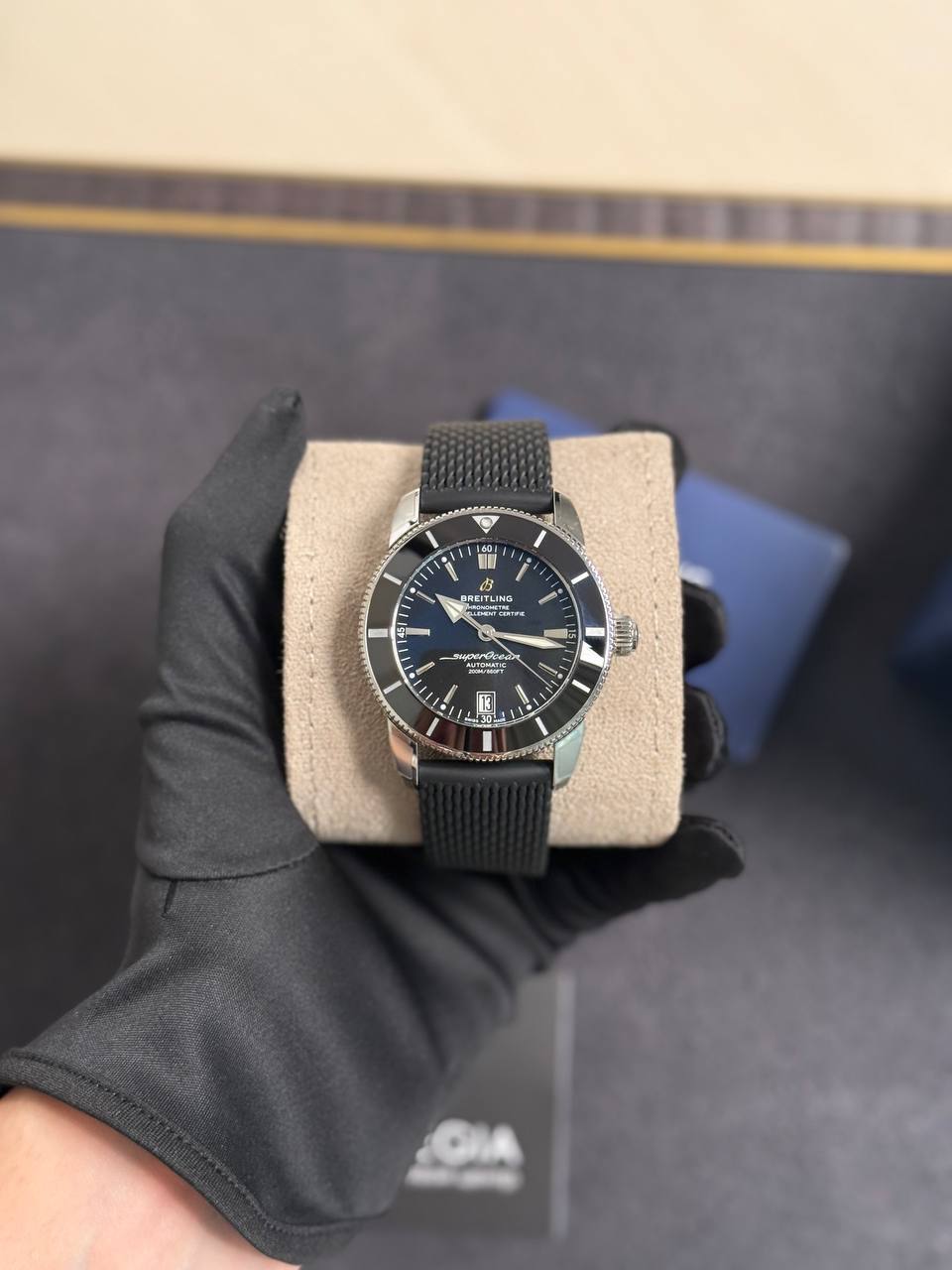 Breitling Superocean automatic wristwatch with black dial and bezel on a beige display cushion, held by a gloved hand.