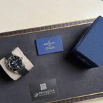 Breitling wristwatch with black dial on a beige cushion beside a blue warranty card and navy box on a dark display mat.