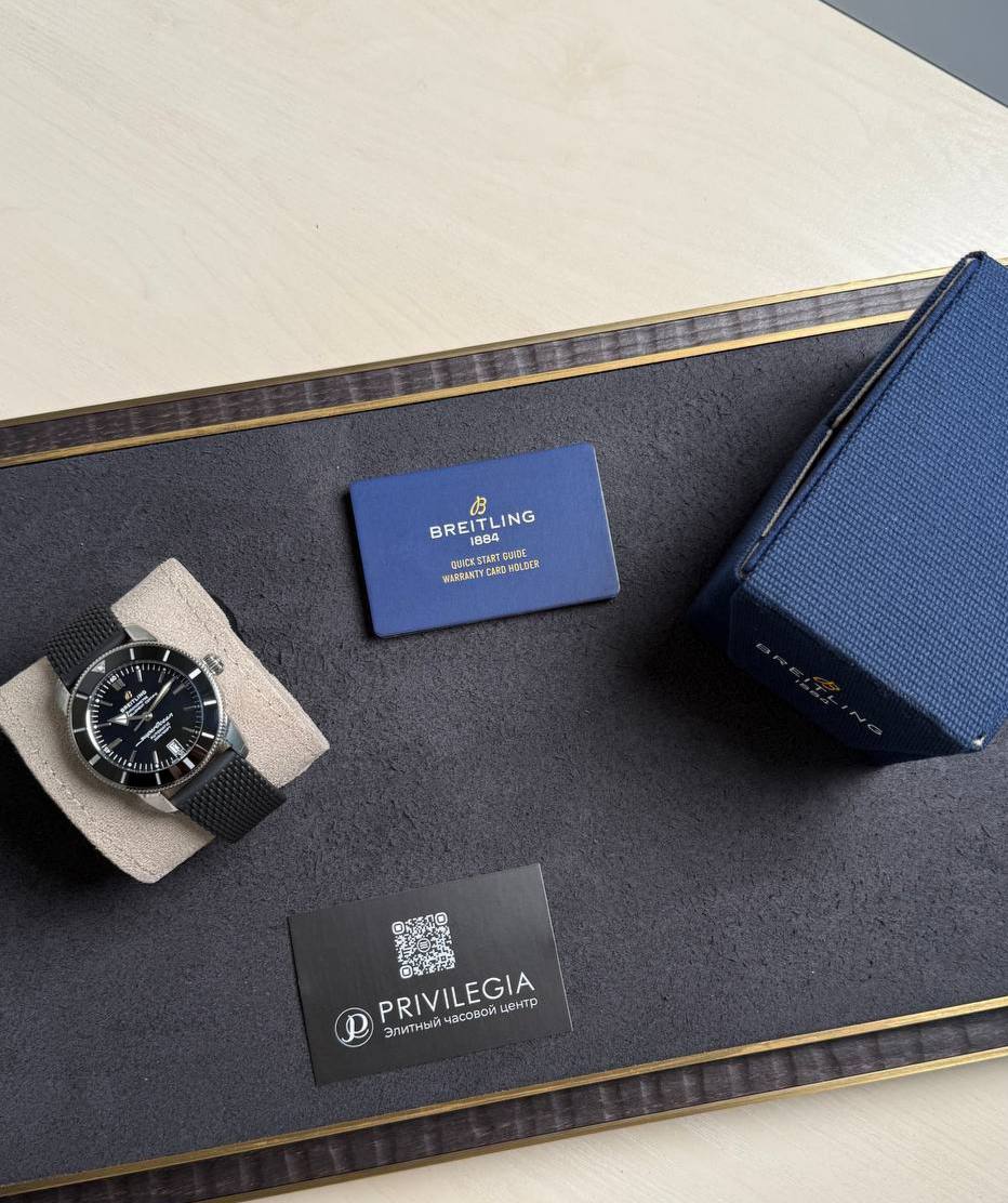 Breitling wristwatch with black dial on a beige cushion beside a blue warranty card and navy box on a dark display mat.
