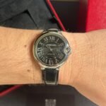 Cartier wristwatch with a black dial, Roman numerals, silver case, and black leather strap on a hairy wrist.