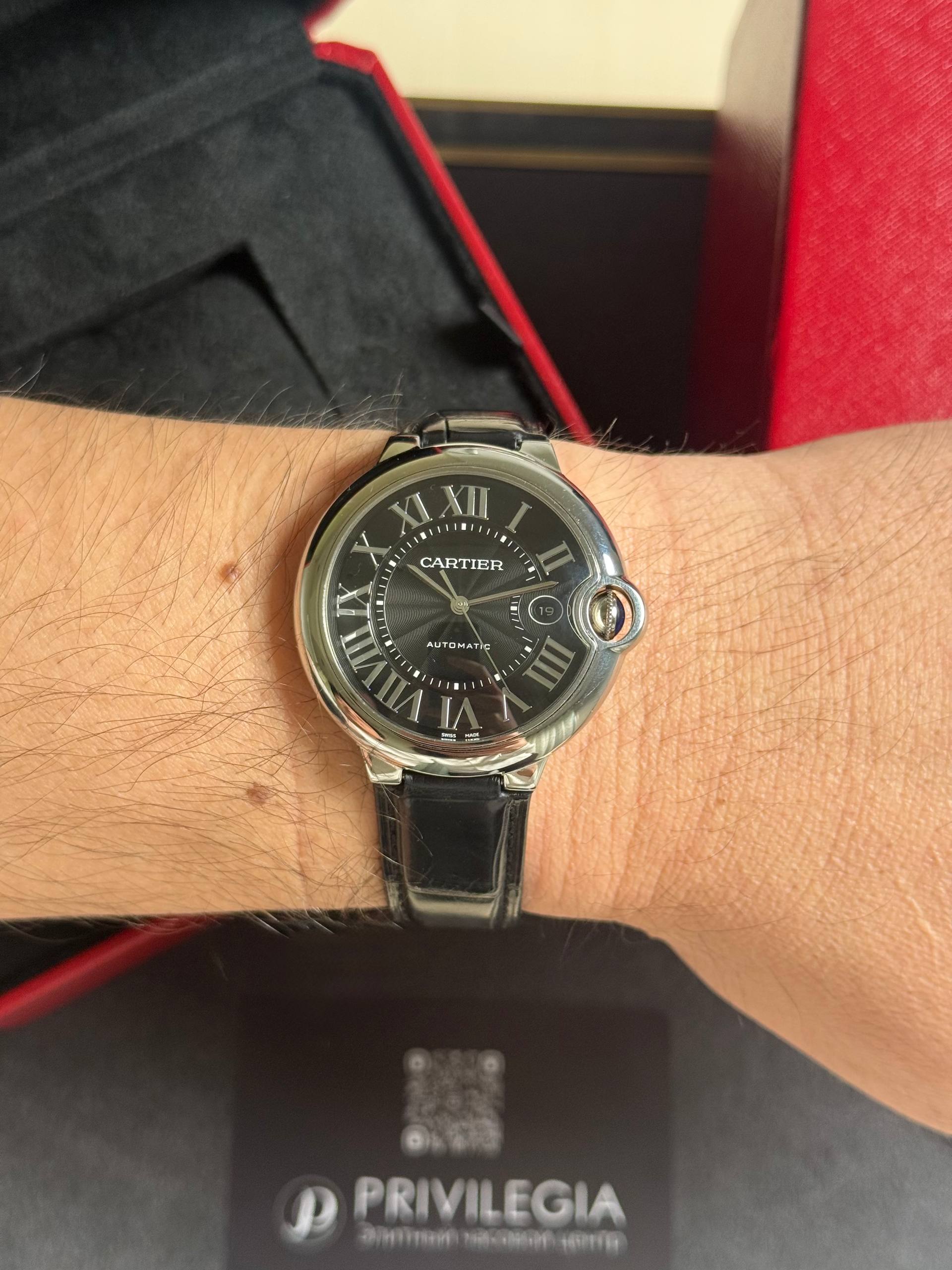 Cartier wristwatch with a black dial, Roman numerals, silver case, and black leather strap on a hairy wrist.
