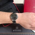 Woman's wrist wearing a Cartier black-dial watch with Roman numerals and leather strap, in front of red Cartier box.