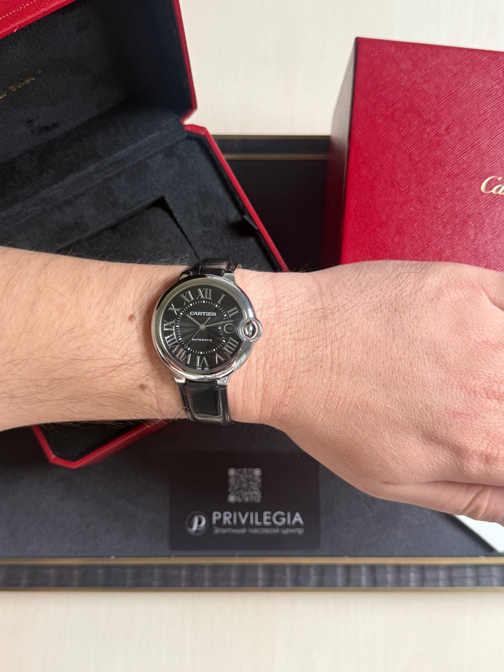 Woman's wrist wearing a Cartier black-dial watch with Roman numerals and leather strap, in front of red Cartier box.