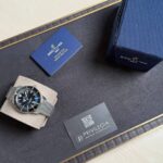 Luxury Breitling watch with metal mesh band displayed on a cushioned stand beside a blue box and business card—watch presentation set.",