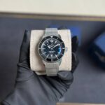 Hand wearing black glove holds a silver mesh-band Breitling SuperOcean wristwatch on a beige cushion, with a dark dial and rotating bezel.