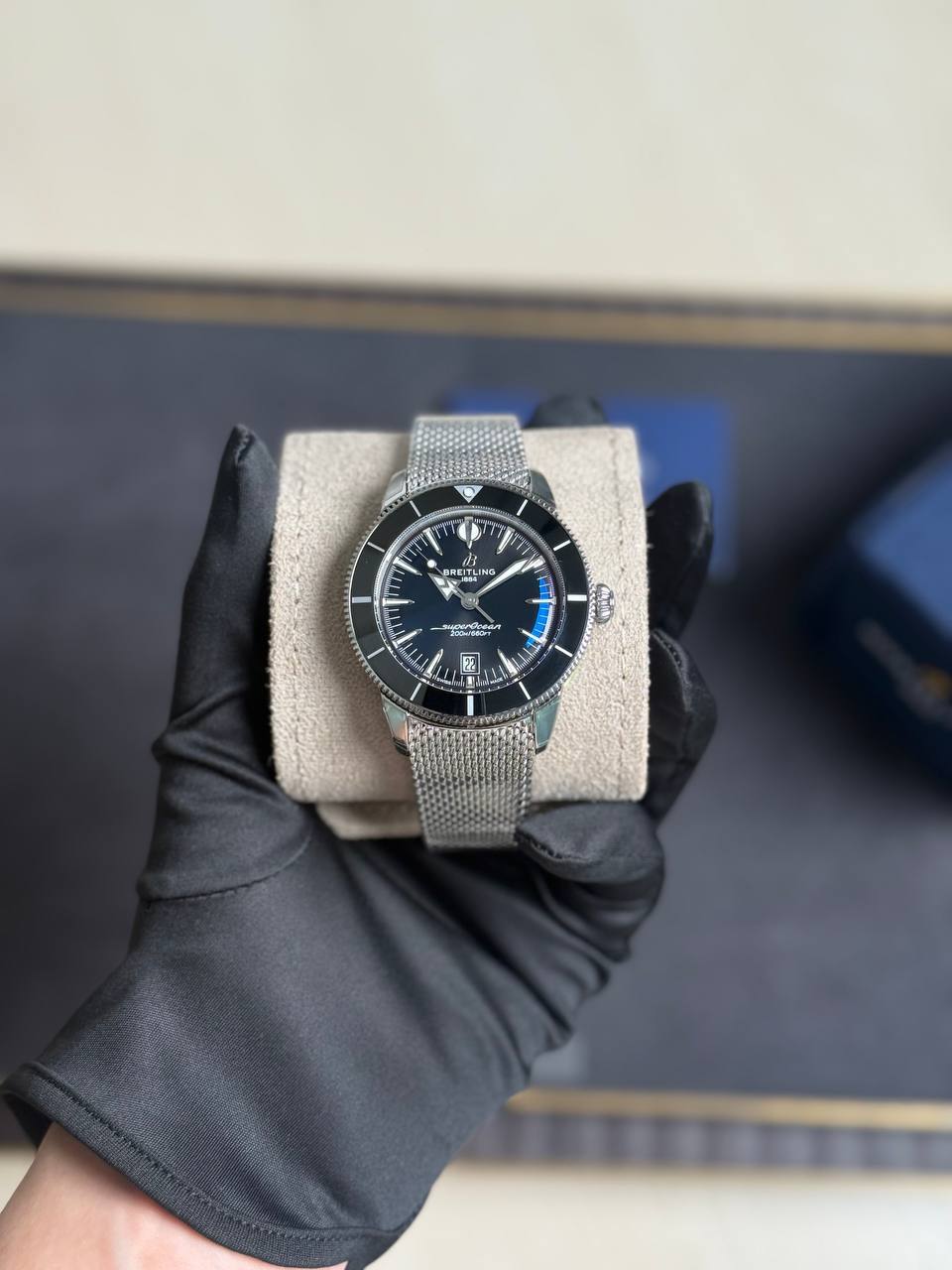 Hand wearing black glove holds a silver mesh-band Breitling SuperOcean wristwatch on a beige cushion, with a dark dial and rotating bezel.