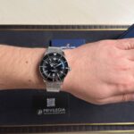 Breitling watch on a wrist with a black bezel and silver mesh bracelet, pictured on a dark box backdrop.