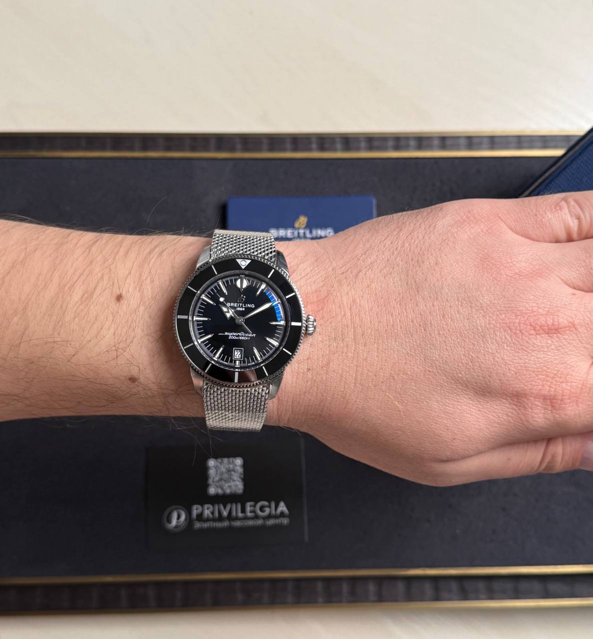 Breitling watch on a wrist with a black bezel and silver mesh bracelet, pictured on a dark box backdrop.