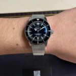 Wrist wearing a black Breitling Superocean diver's watch with a silver mesh band and bezel, shown in close-up with a Privilega card in the background.