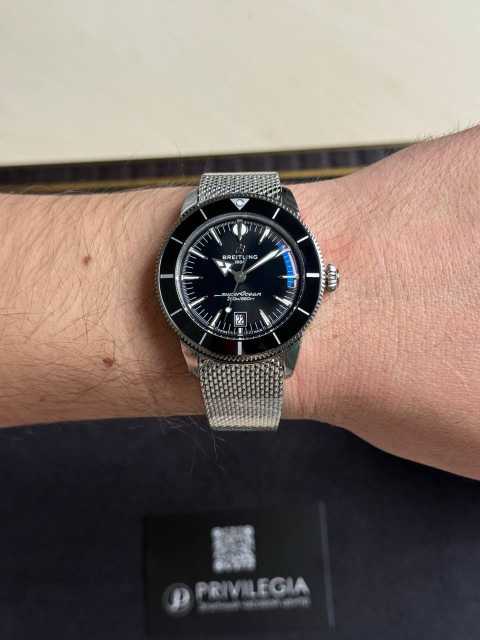 Wrist wearing a black Breitling Superocean diver's watch with a silver mesh band and bezel, shown in close-up with a Privilega card in the background.