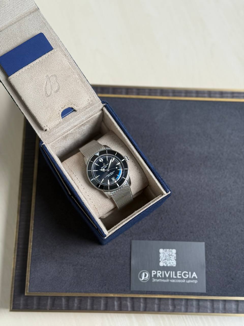 Silver mesh-band wristwatch in an open beige presentation box inside a blue outer box; Privilegia card visible nearby.