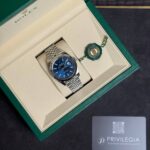 Rolex watch in its green presentation box with a blue dial and a linked metal bracelet, tag attached nearby.