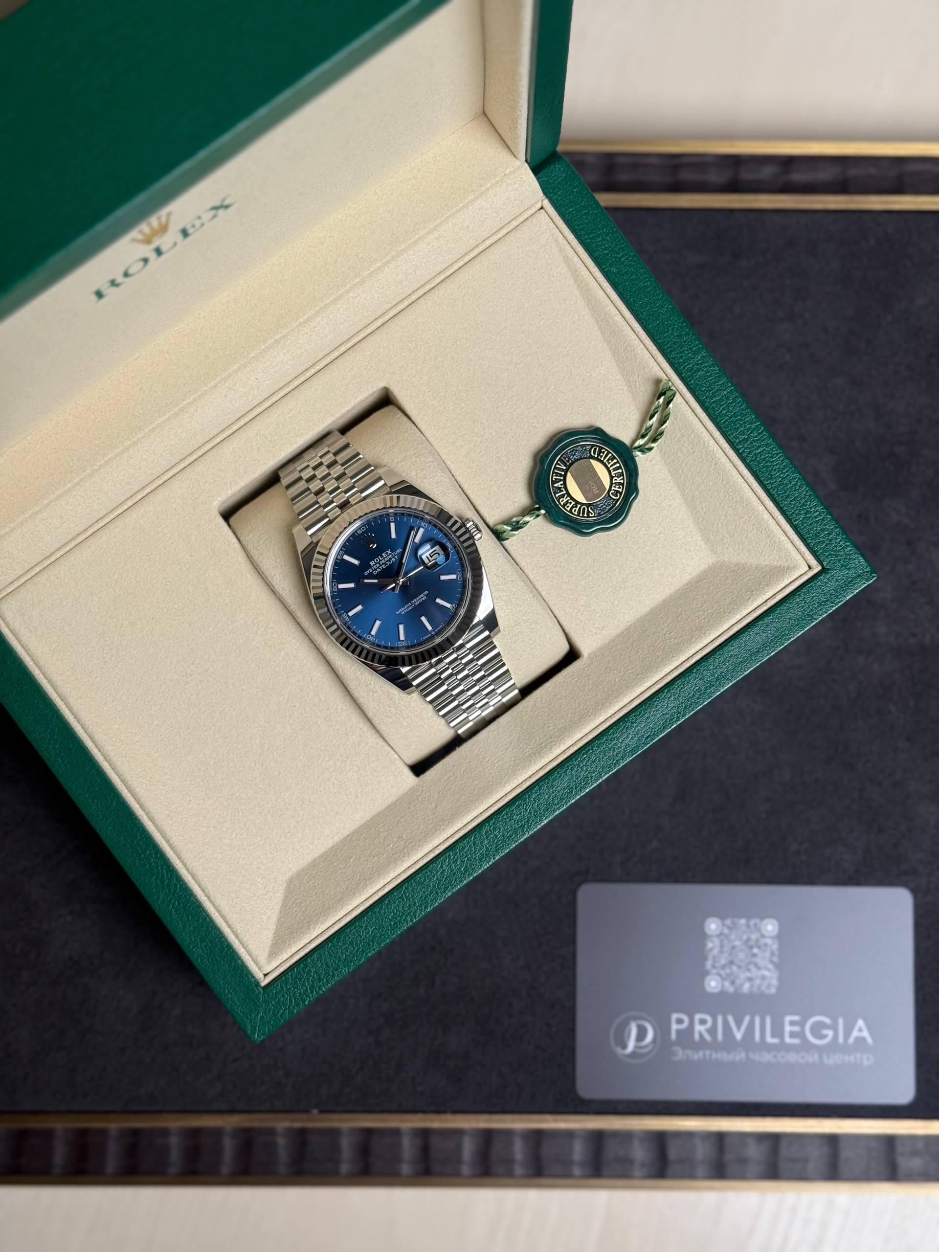 Rolex watch in its green presentation box with a blue dial and a linked metal bracelet, tag attached nearby.