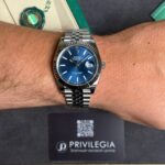 Silver Rolex Datejust on a metal bracelet with a blue dial and date window at 3 o'clock on a wrist; green tag and Privilegia card in background.