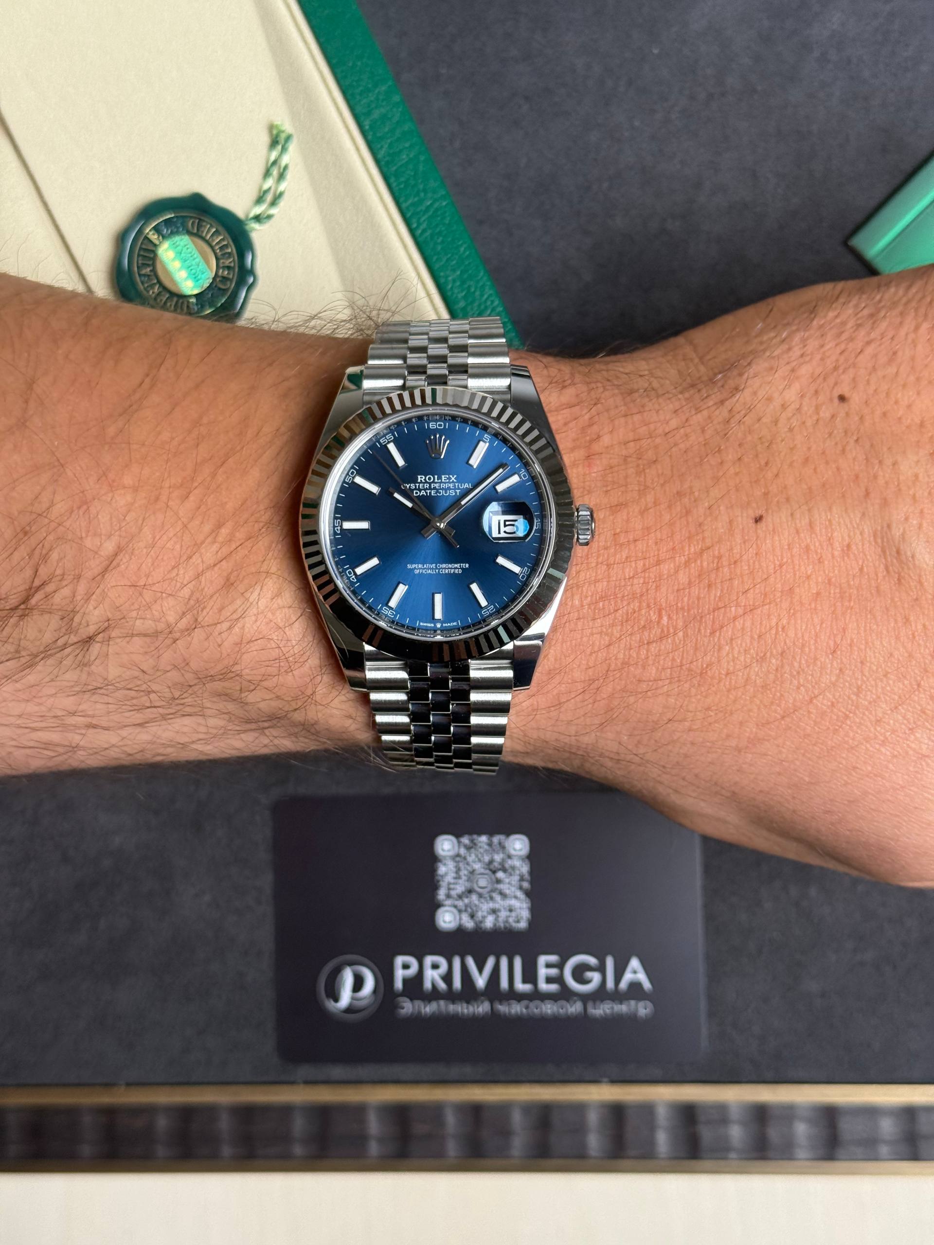Silver Rolex Datejust on a metal bracelet with a blue dial and date window at 3 o'clock on a wrist; green tag and Privilegia card in background.