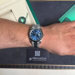 Wrist wearing a silver Rolex with blue dial, over a black display mat and a Privilegia card in the background.