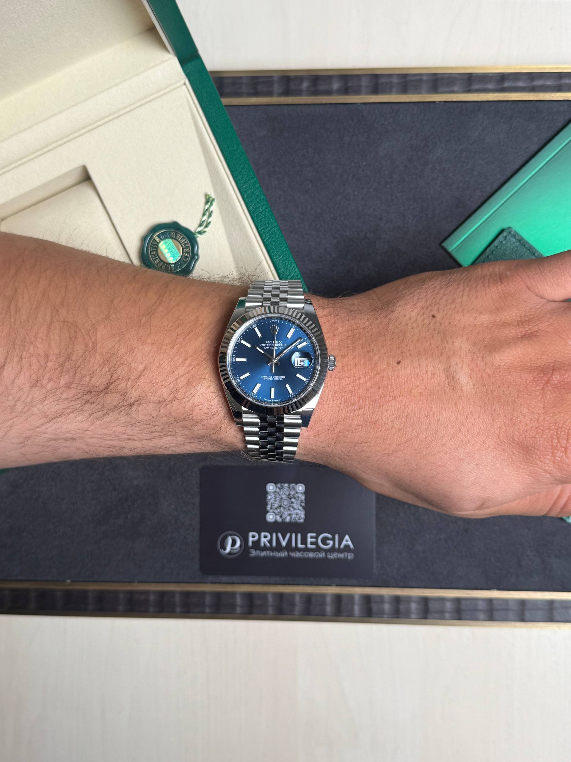 Wrist wearing a silver Rolex with blue dial, over a black display mat and a Privilegia card in the background.