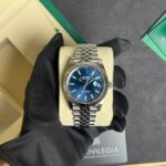 Rolex Datejust wristwatch with a blue dial, fluted bezel, and stainless steel Jubilee bracelet held by a gloved hand on a display cushion.