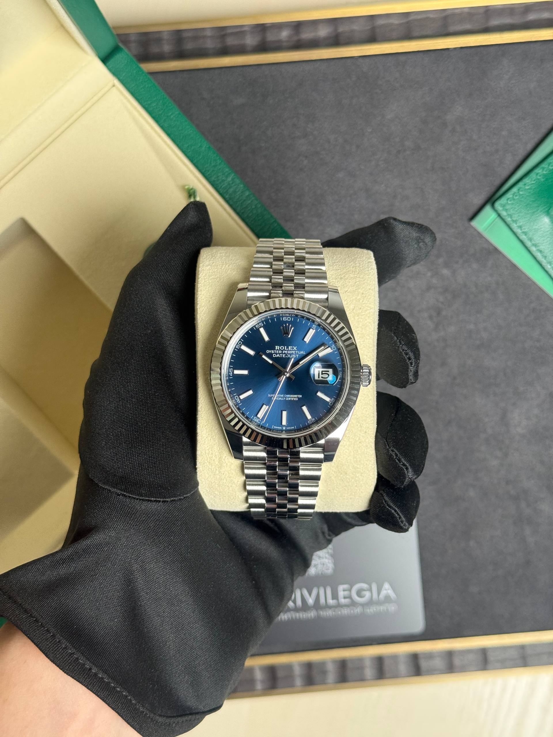 Rolex Datejust wristwatch with a blue dial, fluted bezel, and stainless steel Jubilee bracelet held by a gloved hand on a display cushion.