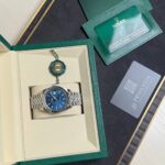 Rolex watch with blue dial in beige presentation box, green outer case and accompanying warranty card and seals nearby.
