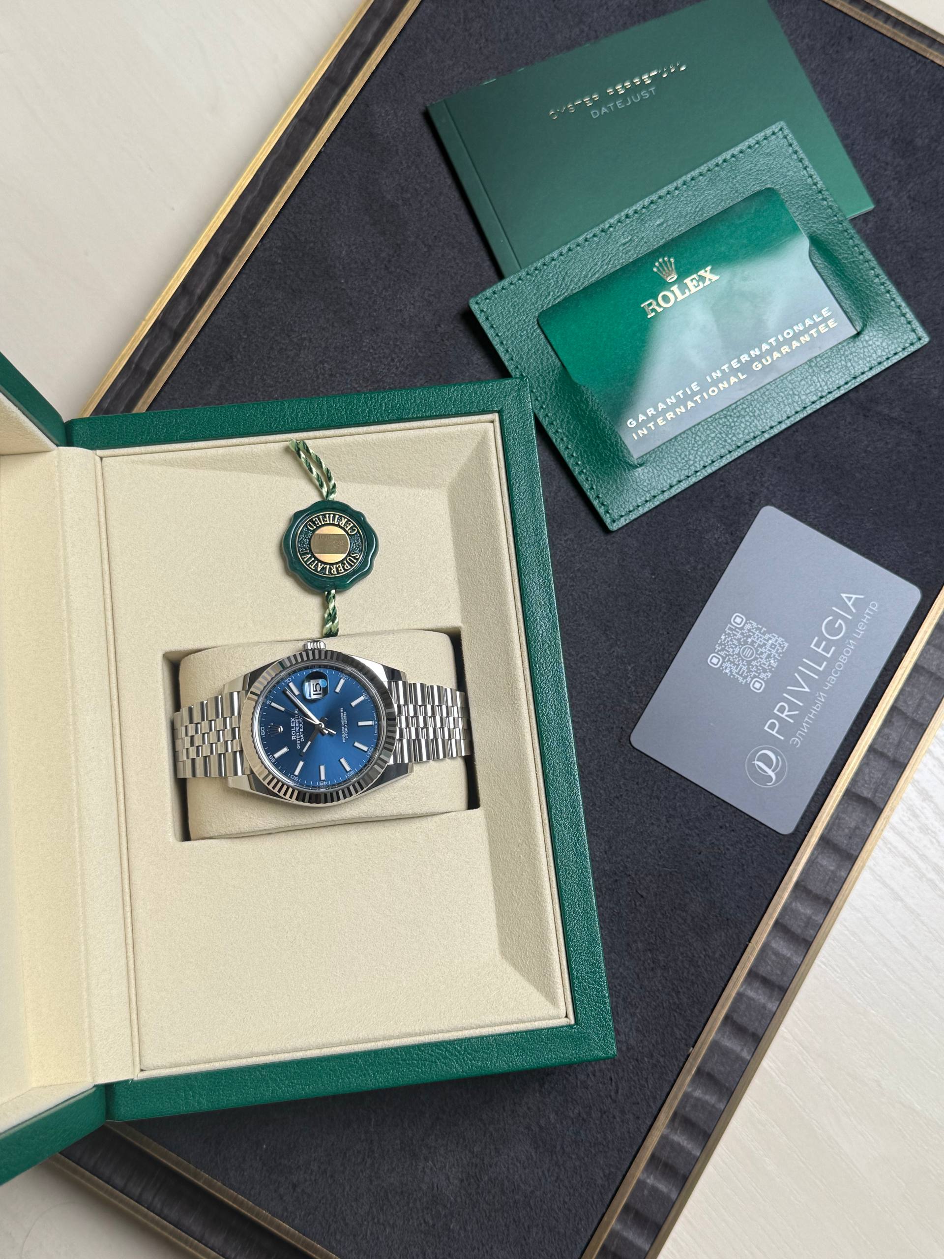 Rolex watch with blue dial in beige presentation box, green outer case and accompanying warranty card and seals nearby.