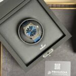Hublot wristwatch with a blue strap sitting in a black display box with the brand logo nearby.