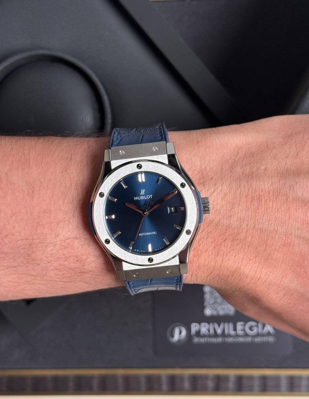 Hublot automatic wristwatch with blue sunburst dial, date window, silver bezel, and blue strap on the wrist.