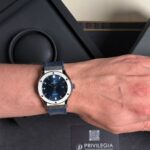 Blue-dial Hublot automatic watch with a silver bezel and blue leather strap on a wrist, shown in a luxury box setting in the background.