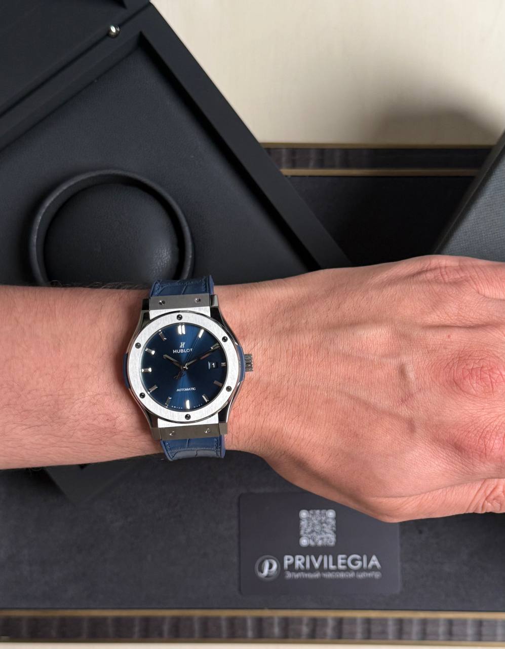 Blue-dial Hublot automatic watch with a silver bezel and blue leather strap on a wrist, shown in a luxury box setting in the background.