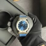 Close-up of a blue-dial Hublot wristwatch with a silver case and blue strap being held by a gloved hand. It’s an automatic watch with a date window on the right side.