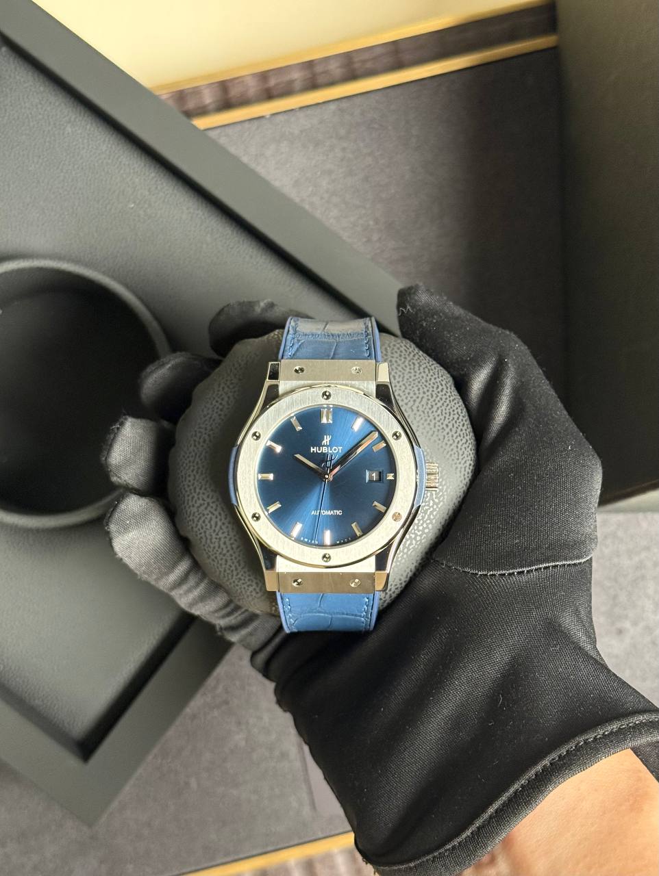 Close-up of a blue-dial Hublot wristwatch with a silver case and blue strap being held by a gloved hand. It’s an automatic watch with a date window on the right side.