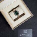 Luxury wristwatch with emerald-green dial and diamond-studded bezel displayed in a cream presentation box; two-tone bracelet shows a polished finish.