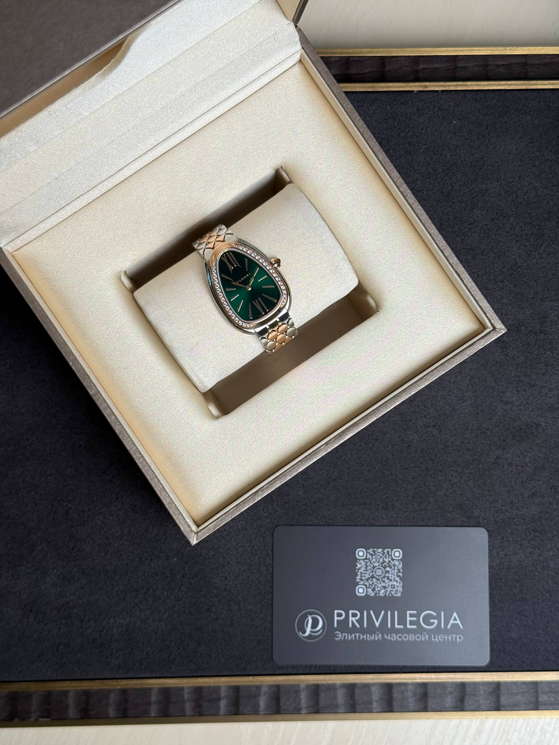 Luxury wristwatch with emerald-green dial and diamond-studded bezel displayed in a cream presentation box; two-tone bracelet shows a polished finish.