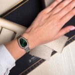 Wrist wearing a silver metal bracelet watch with a green dial and jeweled bezel, resting on fabric swatches in a presentation box
