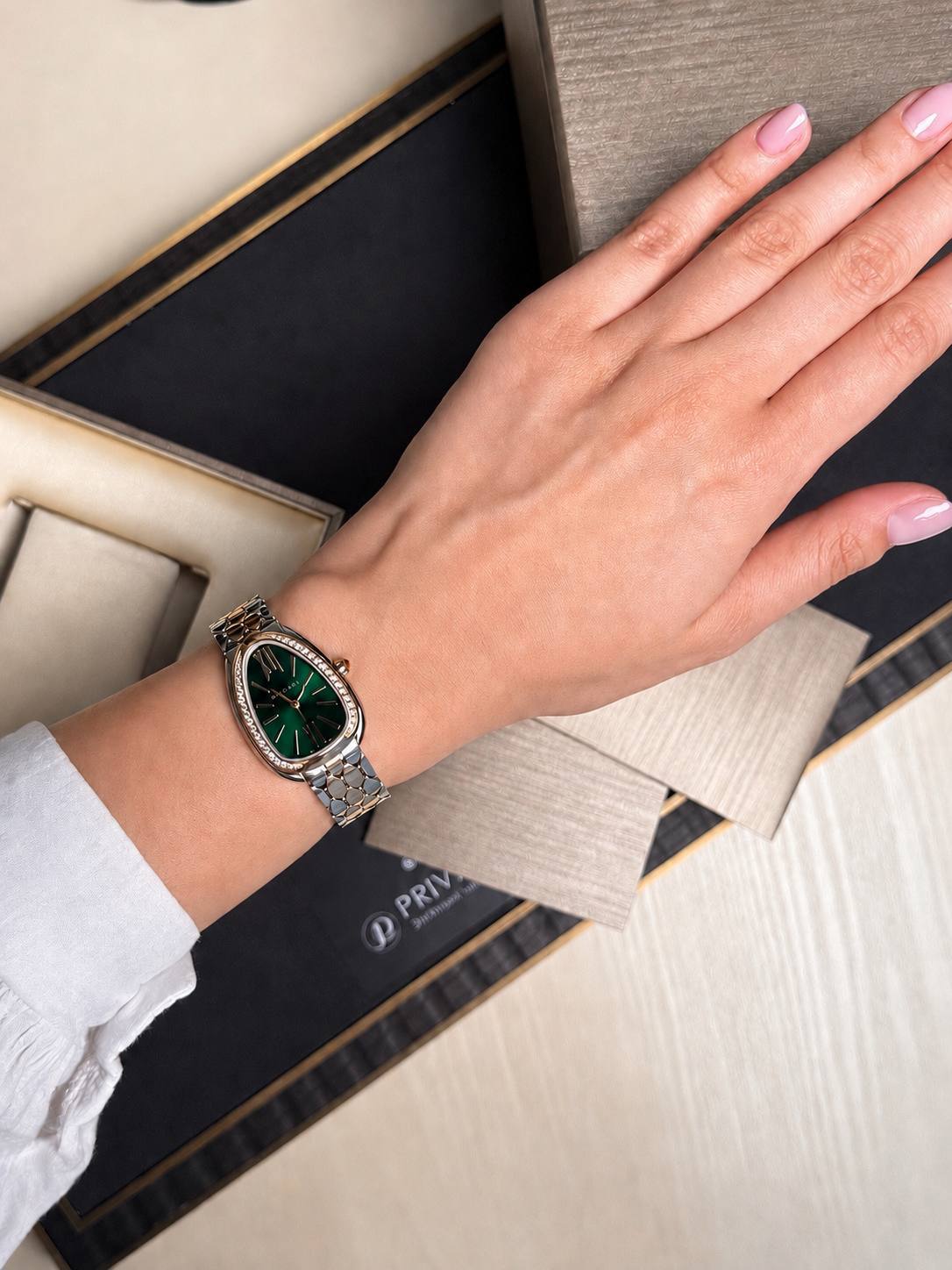 Wrist wearing a silver metal bracelet watch with a green dial and jeweled bezel, resting on fabric swatches in a presentation box