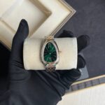 Two-tone luxury wristwatch with a green dial and diamond-studded bezel held by a black-gloved hand over a cushioned display, with an open box in the background.