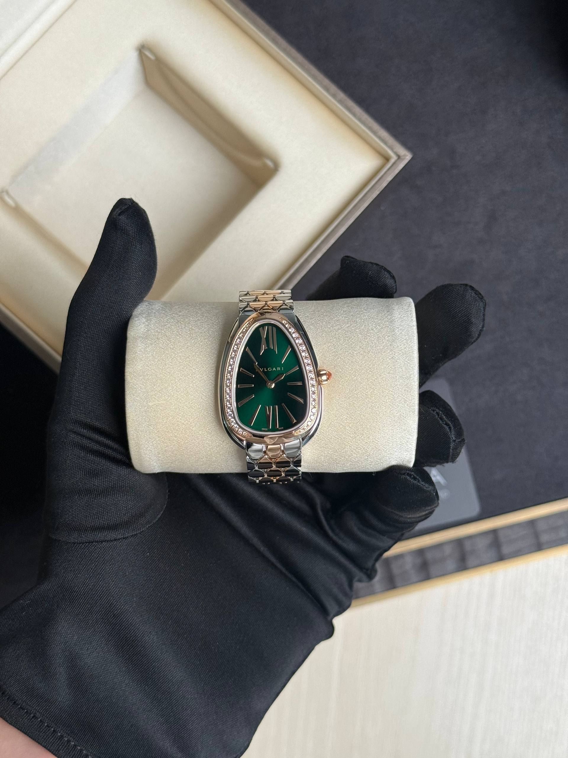 Two-tone luxury wristwatch with a green dial and diamond-studded bezel held by a black-gloved hand over a cushioned display, with an open box in the background.