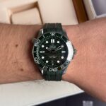 Wrist wearing a dark green Omega Seamaster diver's watch with a black dial and white markers, on a green strap.