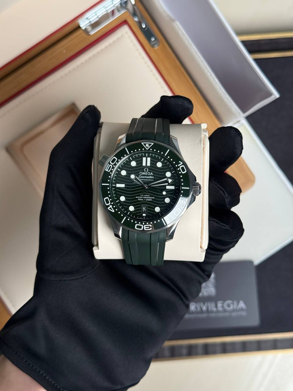 Green Omega Seamaster wristwatch on a cushioned display held by a gloved hand in a box.