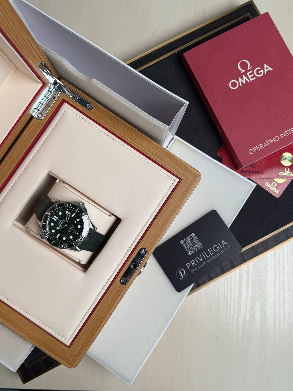 Omega wristwatch in a beige leather display box with a wooden frame and red interior lining, along with accompanying manuals and Privilege card nearby