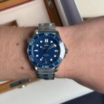 Close-up of a blue Omega Seamaster dive watch with a stainless steel bracelet worn on a forearm. The watch features a blue bezel, large luminous markers, and a date window at 6 o'clock.