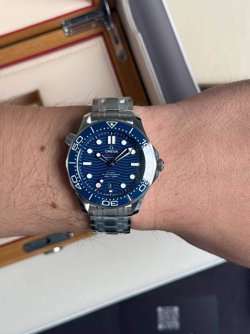 Close-up of a blue Omega Seamaster dive watch with a stainless steel bracelet worn on a forearm. The watch features a blue bezel, large luminous markers, and a date window at 6 o'clock.