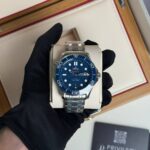 Omega dive watch with blue bezel and dial in a stainless-steel bracelet, held by a gloved hand over a white display cushion