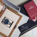 Omega wristwatch in a wooden presentation box with cream interior, blue dial, stainless steel bracelet, surrounded by manuals and cards
