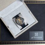 Skeleton-style gold-toned wristwatch with a dark strap in a grey velvet-lined presentation box.
