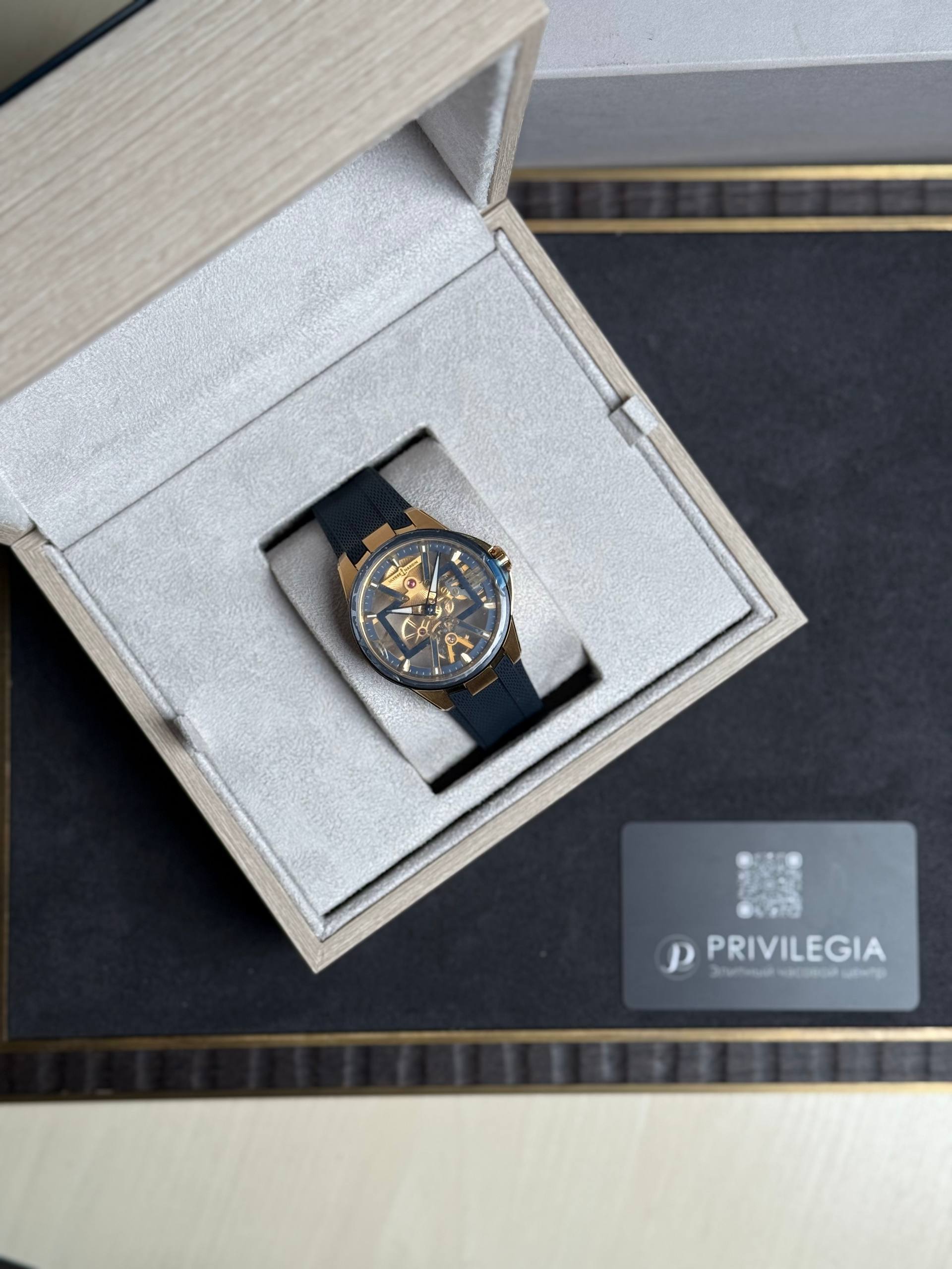Skeleton-style gold-toned wristwatch with a dark strap in a grey velvet-lined presentation box.